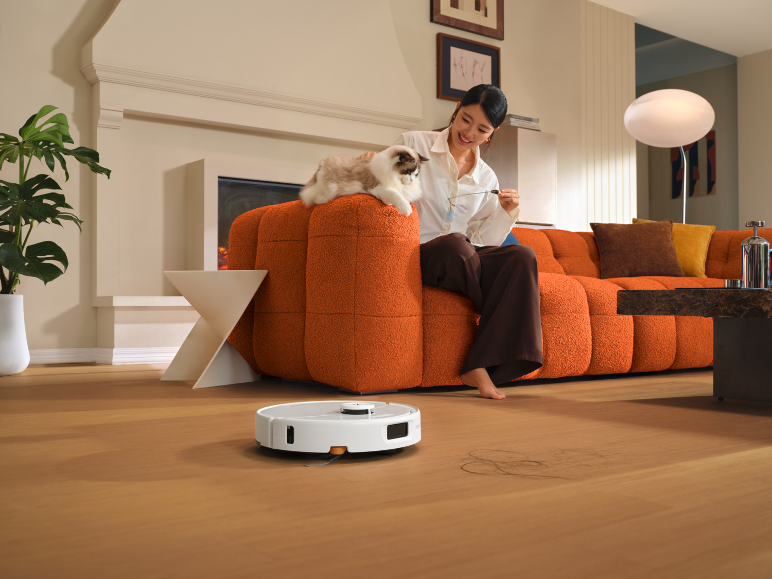 Robotstofzuiger op houten vloer, vrouw met kat op oranje bank.