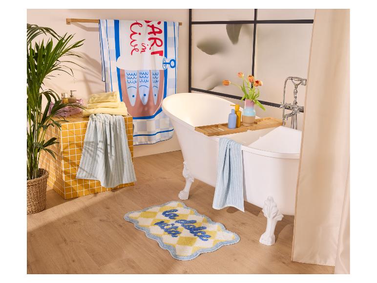 Badkamer met wit bad, gele tegels, plant en handdoeken, met een vloerkleedje met de tekst 'la dolce vita'.