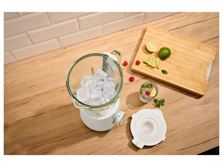 Blender met ijs, limoenen, frambozen en munt op een houten oppervlak.