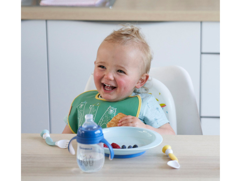 Een baby eet een koekje, met een  'Babymoov' flesje, een bordje en bestek op tafel.
