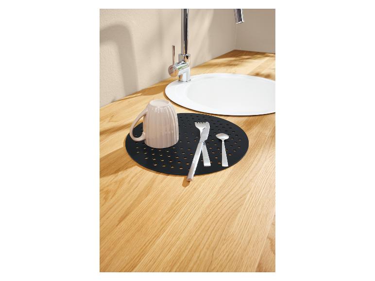 Een zwarte, geperforeerde droogmat op een licht houten aanrecht, met een mok en bestek bij een witte gootsteen.