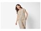 Vrouw in beige overhemd met korte mouwen en broek, lachend op witte achtergrond.