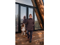 Vrouw in bruine jas en grijze broek met hond op houten terras.