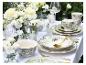 Tafel gedekt met groen en wit bloemig servies, gouden bestek en witte bloemstukken.