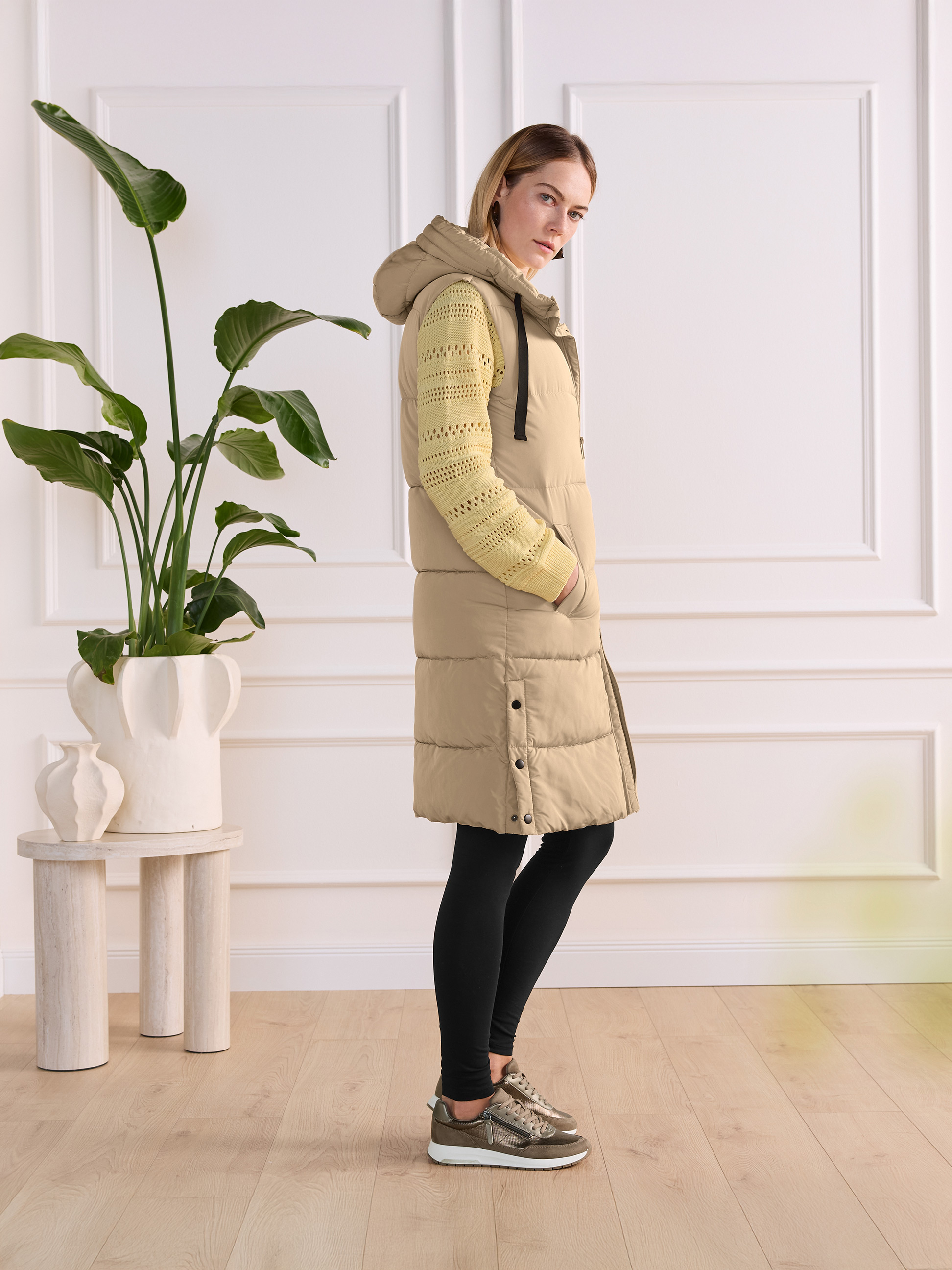 Vrouw in lange beige bodywarmer, gele gebreide trui en zwarte legging met sneakers.