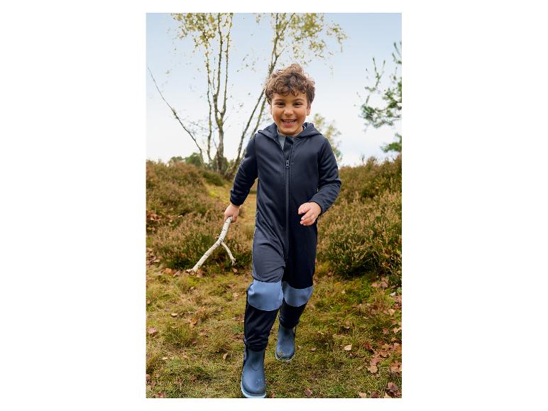 Lachende jongen in een donkerblauwe waterdichte overall en laarzen, met een stok in een bos.
