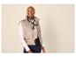 Vrouw in een beige gewatteerd vest, wit shirt met lange mouwen en donkere jeans, lachend.