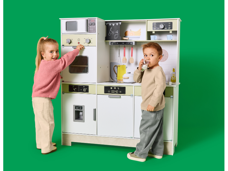 Twee kinderen spelen met een grote houten speelgoedkeuken tegen een groene achtergrond.