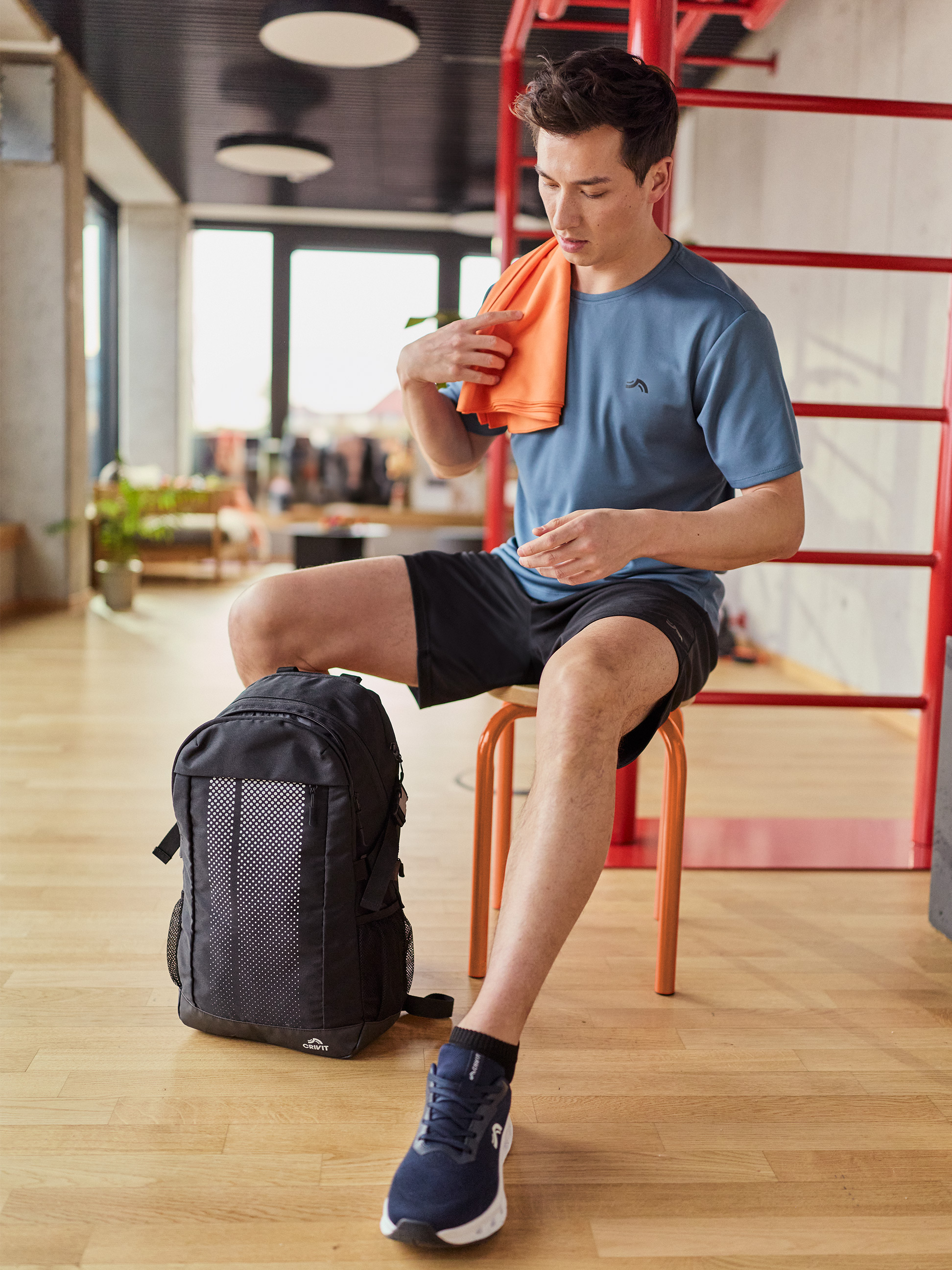 Man in sport-T-shirt en korte broek, droogt zich af met een oranje handdoek, met een rugzak ernaast.