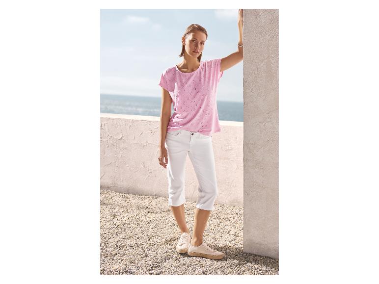 Vrouw in een roze geperforeerd T-shirt en witte capri broek, leunend tegen een muur met zeezicht.