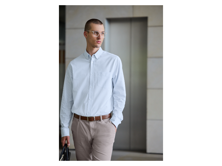Een man in een lichtblauw gestreept overhemd en beige broek draagt een tas.