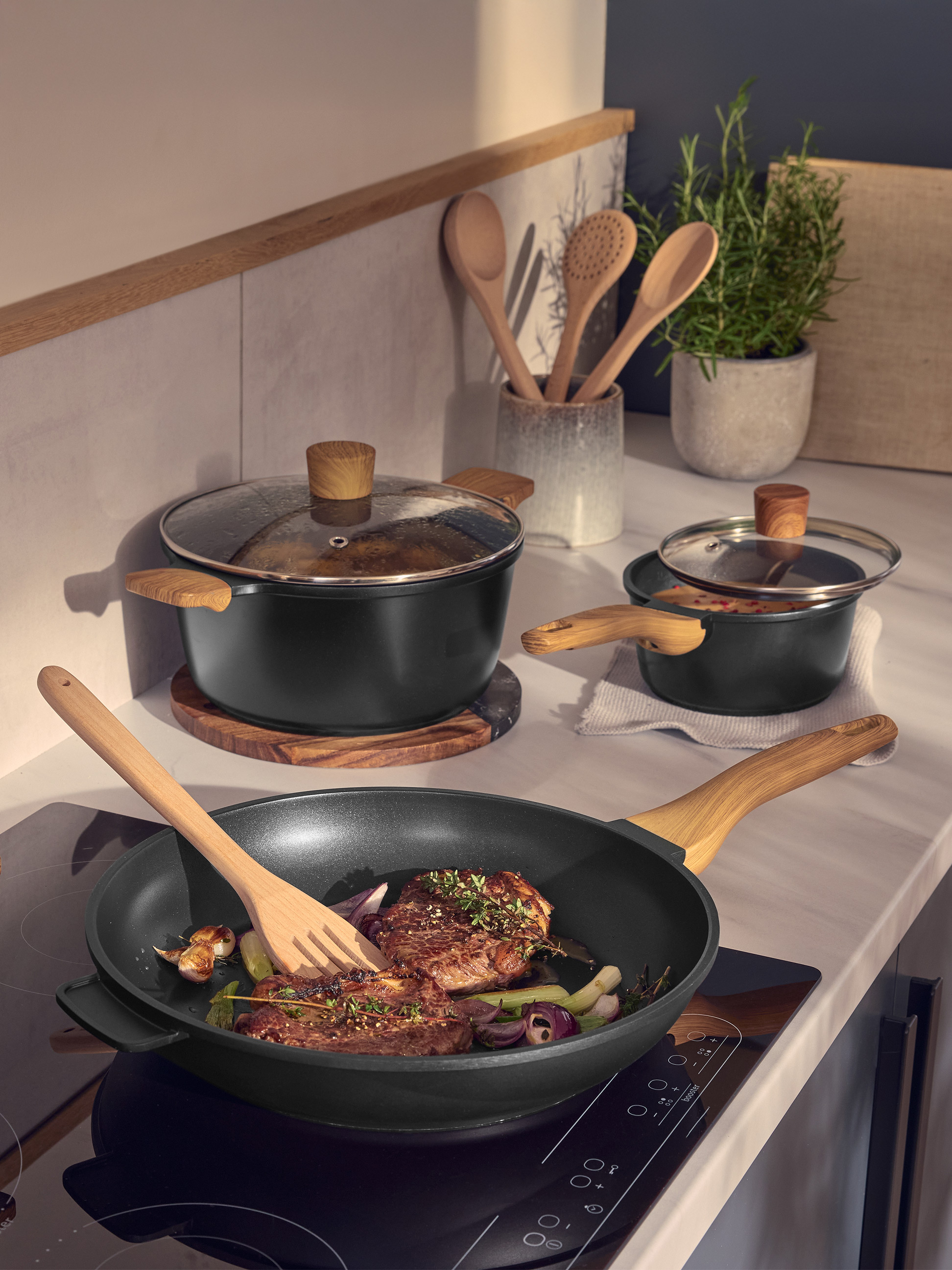 Zwarte pannen en potten met houten handvatten, vlees en groenten koken in een moderne keuken.