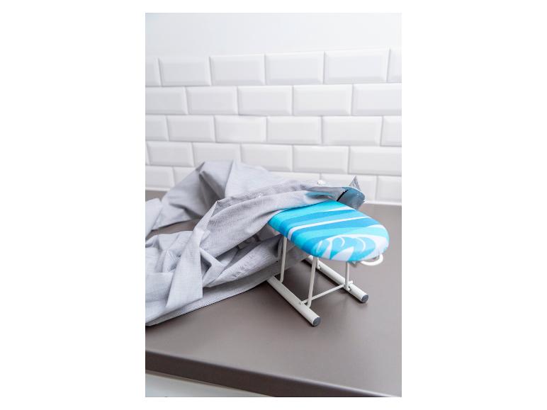 Een mini strijkplank met een blauw-wit gestreept hoesje staat op een grijs aanrecht met een grijs overhemd erop.