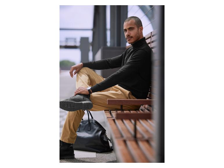 Een man zit op een bank met een zwarte trui, beige broek en zwarte laarzen.