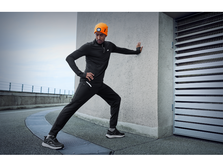 Man in zwarte hardloopoutfit en oranje muts met hoofdlamp rekt zich uit tegen een muur.