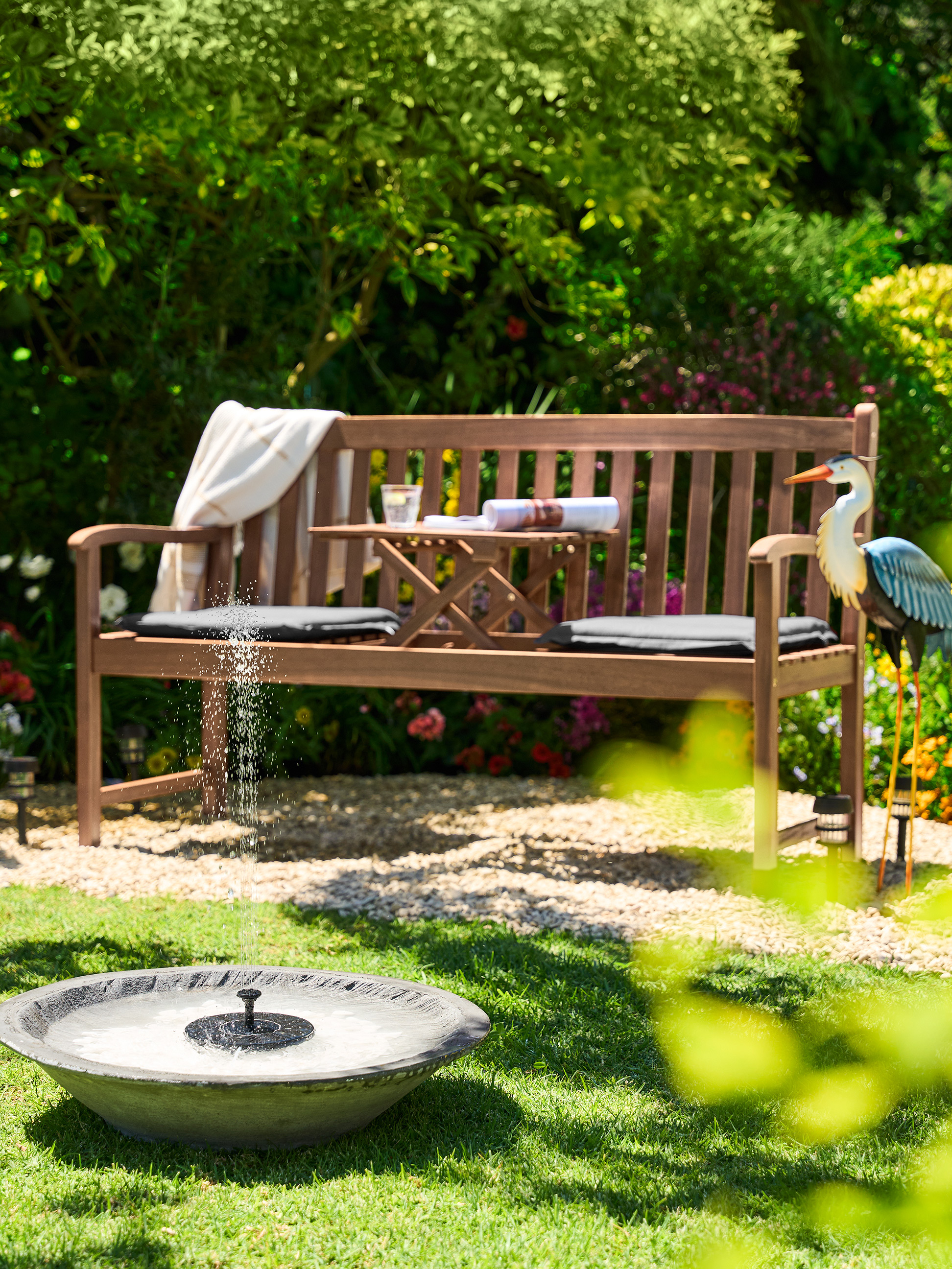 Houten tuinbank met een tafeltje, kussens en een decoratieve reiger in een zonnige tuin.