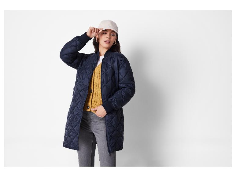 Vrouw in donkerblauwe gewatteerde jas, geel vest, grijze jeans en beige baseballpet.