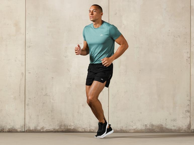 Man in een blauwe T-shirt en zwarte sportbroek, poseert voor een betonnen muur.