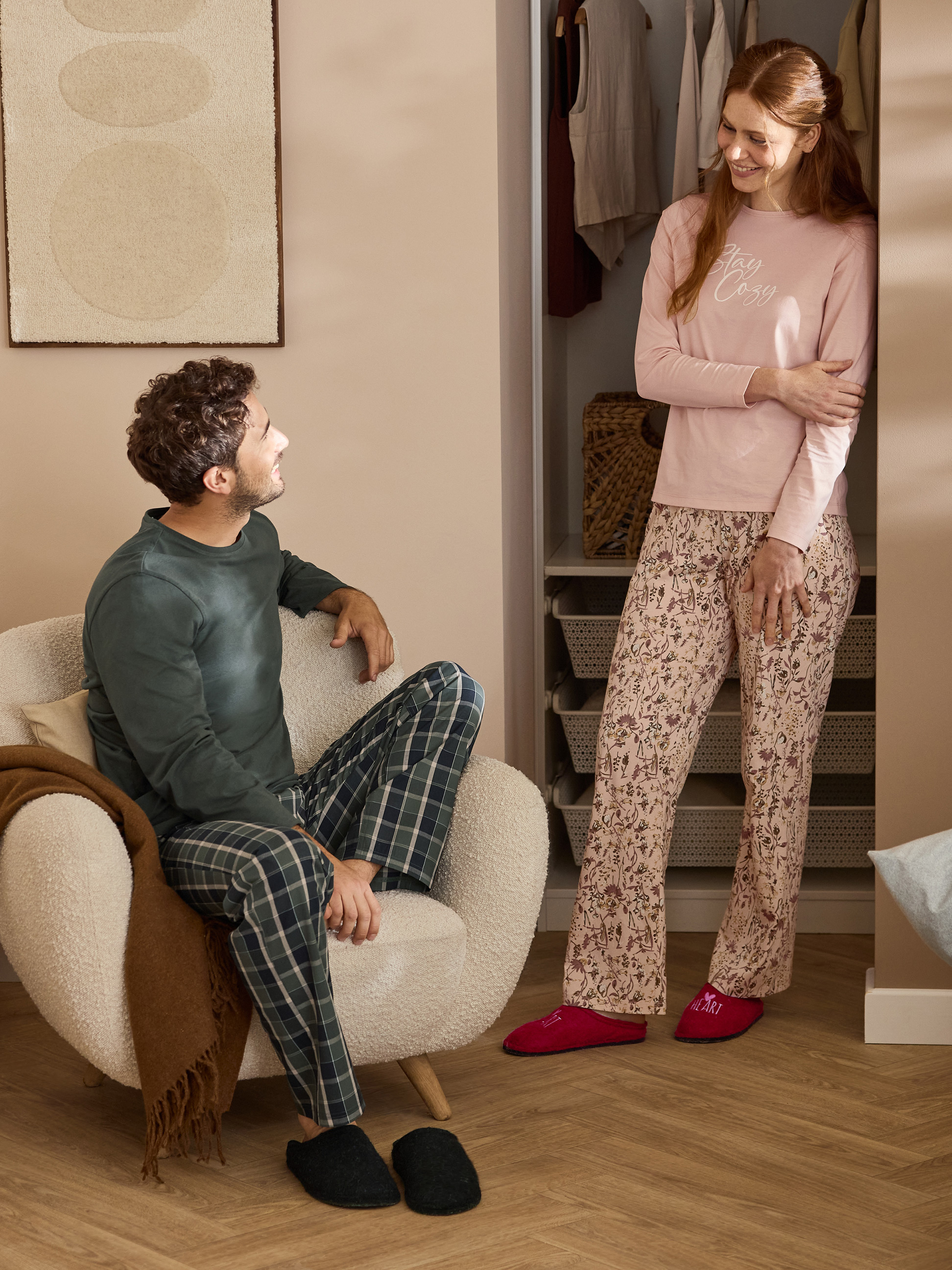 Paar in pyjama en pantoffels, vrouw draagt shirt met tekst „Stay Cozy”.