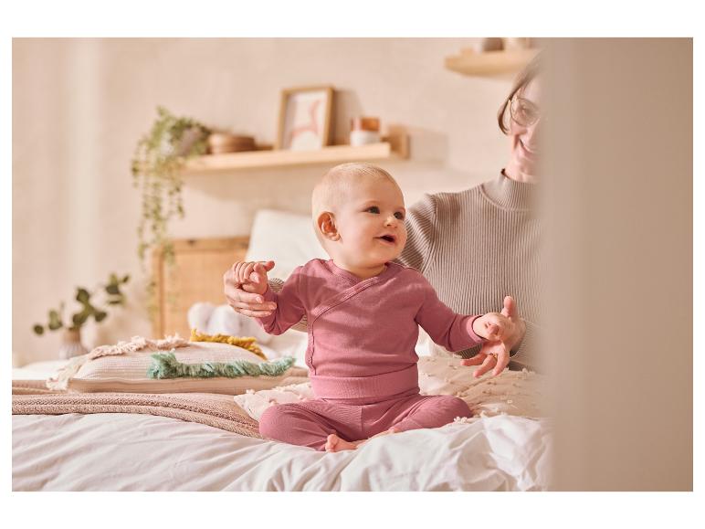Baby in roze kleding zit op een bed met een volwassene.