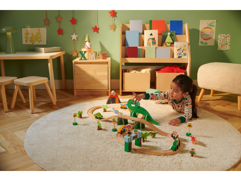 Meisje speelt met houten treinbaan en dinosaurussen op een rond vloerkleed in een kerstkamer.
