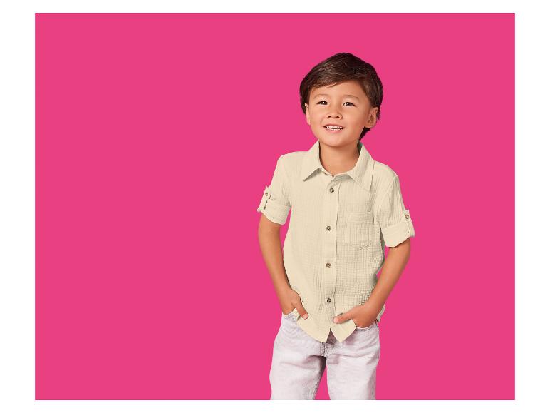 Lachende jongen in een crèmekleurig overhemd en lichte broek tegen een fuchsia achtergrond.
