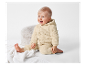 Een blije baby zit op een wit kleed in een beige teddy jumpsuit.