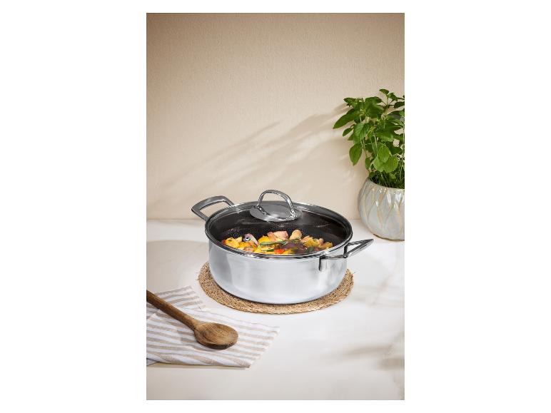 Roestvrijstalen pan met glazen deksel, gevuld met groenten en vlees, op een rieten onderzetter.