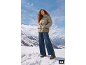 Vrouw in beige pufferjack, wijde jeans en laarzen staand in een besneeuwd berglandschap.