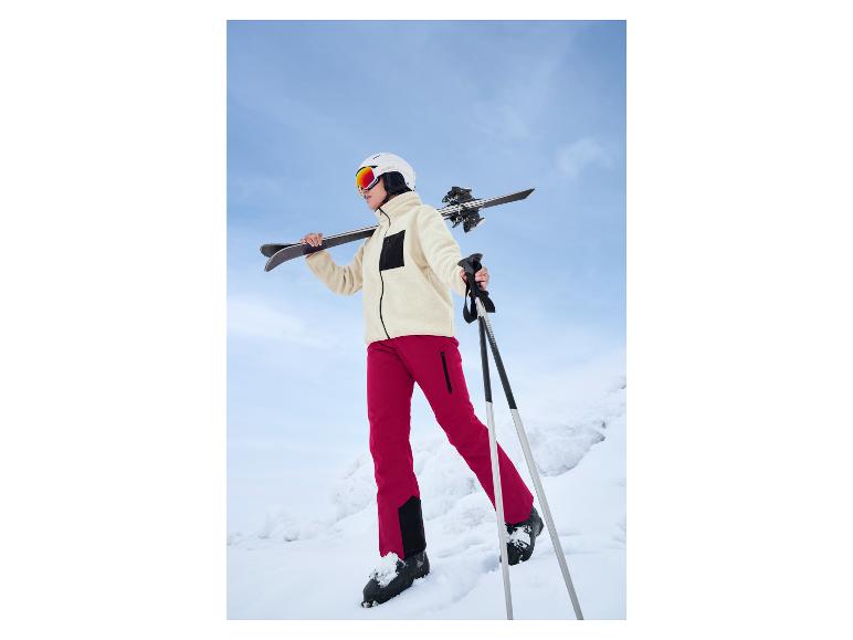 Een vrouw in rode skibroek en een witte Crivit-helm loopt door de sneeuw met ski-uitrusting.