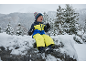 Kind in geel-blauw skipak, muts en handschoenen, zittend op een besneeuwde rots.