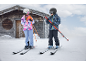 Twee kinderen in skikleding met ski's en stokken in een besneeuwd landschap bij een houten huisje.