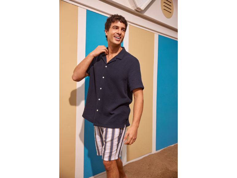 Man in een marineblauw overhemd met korte mouwen en gestreepte shorts, lachend in een strandhuisje.