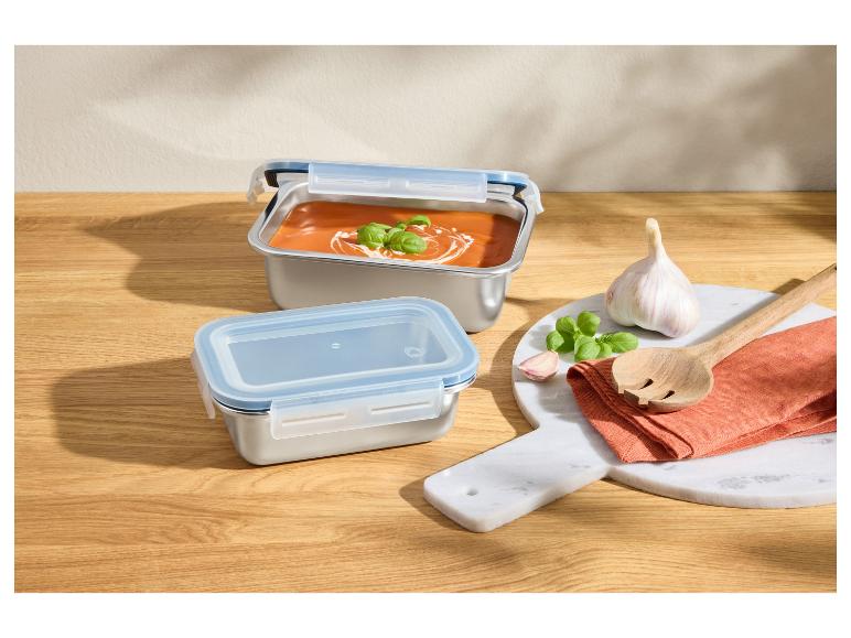 Roestvrijstalen voedselcontainers met tomatensoep, knoflook en basilicum op een houten ondergrond.