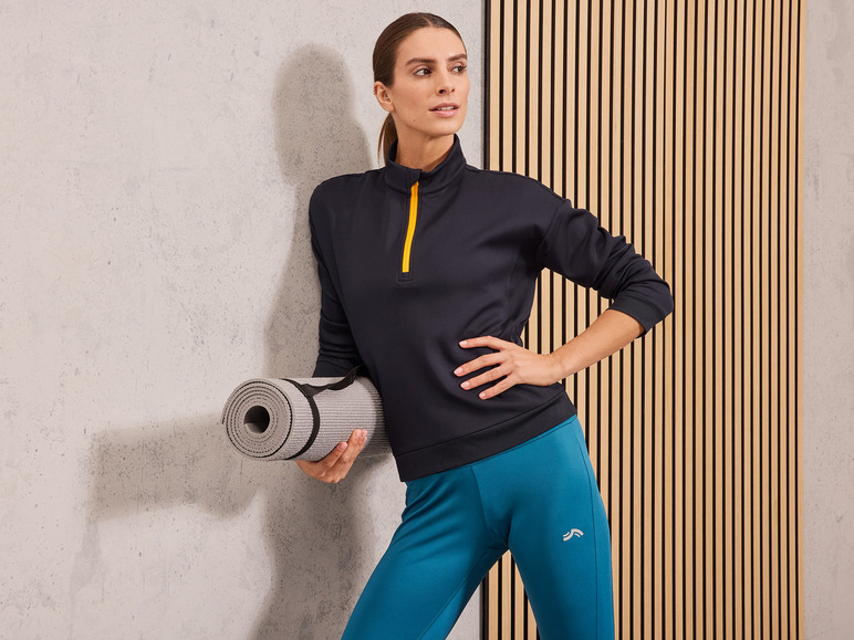 Een vrouw in zwarte sportkleding en blauwe leggings met een yoga mat in haar hand.