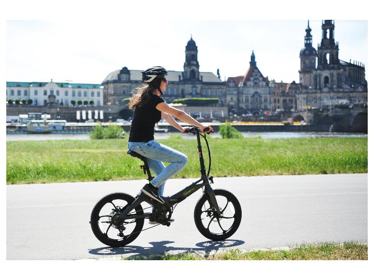 Vrouw fietst op een Sachsengrad e-bike langs een rivier met stadsgezicht op de achtergrond.