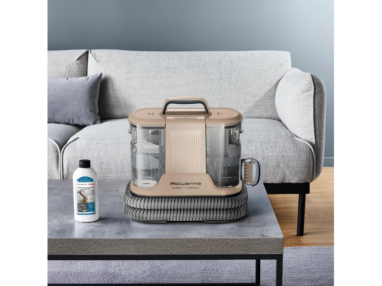 Beige compacte tapijtreiniger op salontafel met reinigingsoplossing en grijze bank.