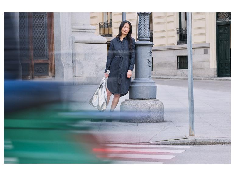 Vrouw in jeansjurk staat bij een lantaarnpaal met een zilveren tas.
