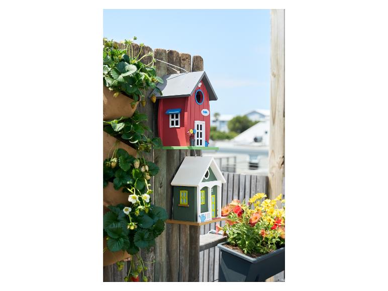 Twee kleurrijke vogelhuisjes bevestigd aan een houten schutting met planten eromheen.