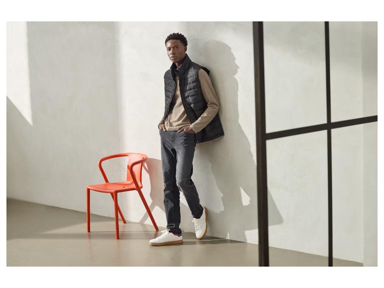 Man in zwart gewatteerd vest, beige shirt met lange mouwen en grijze jeans, leunend tegen een muur.