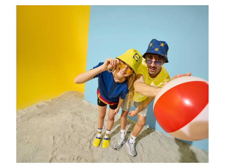Twee personen met bucket hats en zonnebrillen, met een strandbal, tegen een geel-blauwe achtergrond.