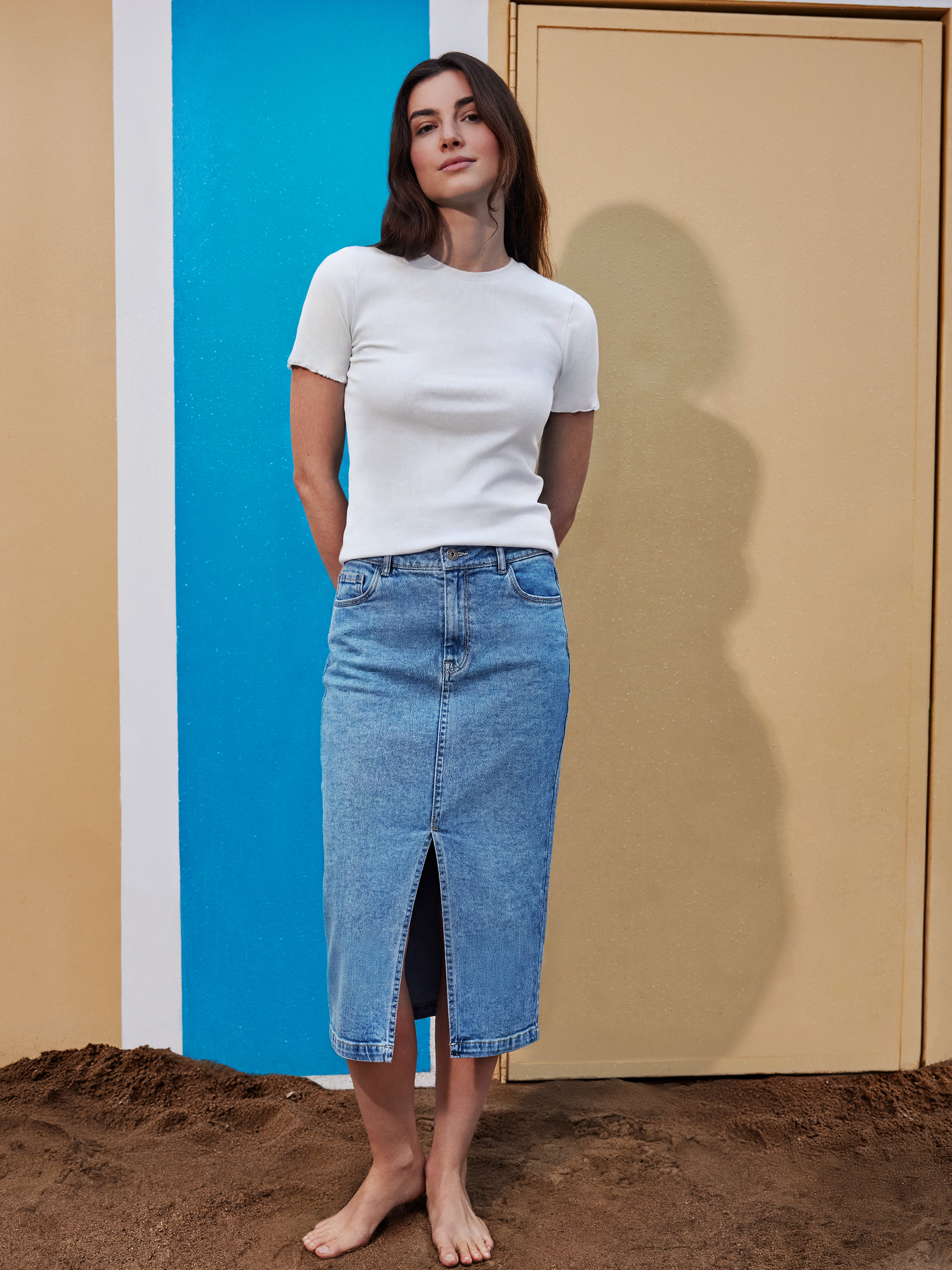 Vrouw in witte top en denim midirok met split, blootsvoets staand in het zand.