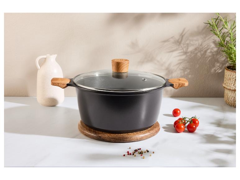 Zwarte pan met houten handvatten en glazen deksel op een houten onderzetter, met tomaten en kruiden.