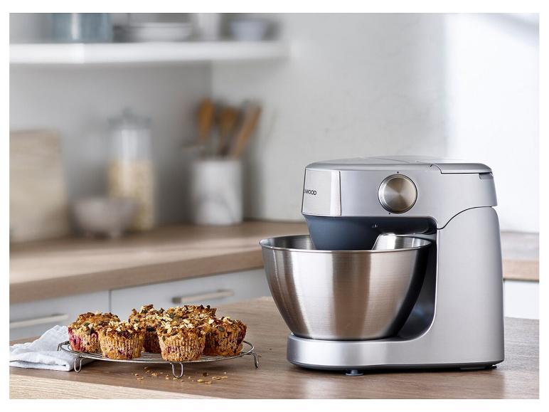 Zilverkleurige keukenmachine met roestvrijstalen kom en muffins op een houten tafel.