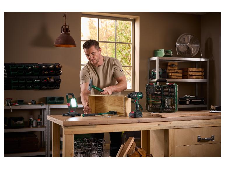 Man monteert een houten kist met een hamer, omringd door elektrisch gereedschap en werkplaatsuitrusting.