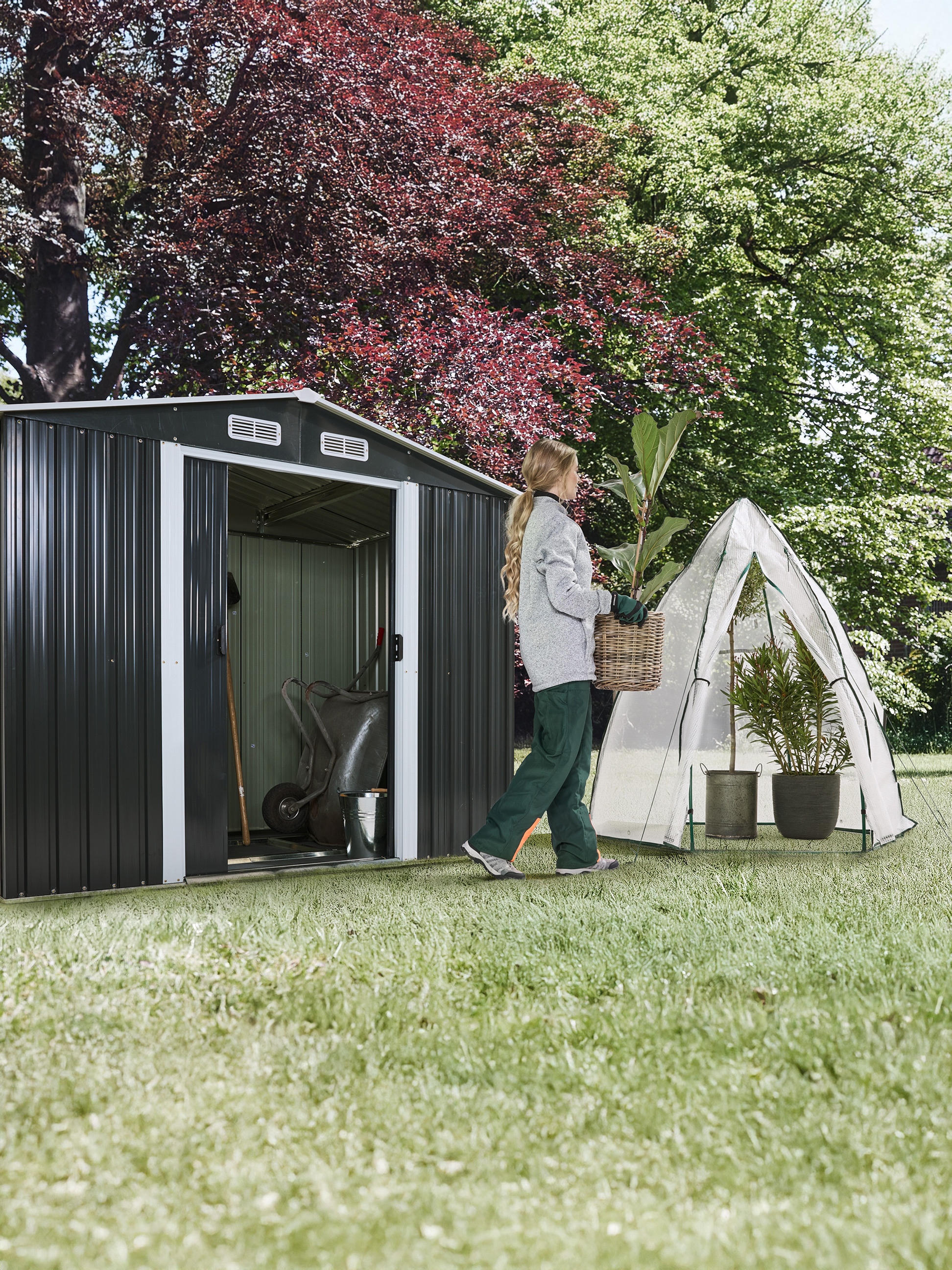 Vrouw draagt plant in een mand, naast een tuinhuisje en een kleine kas.