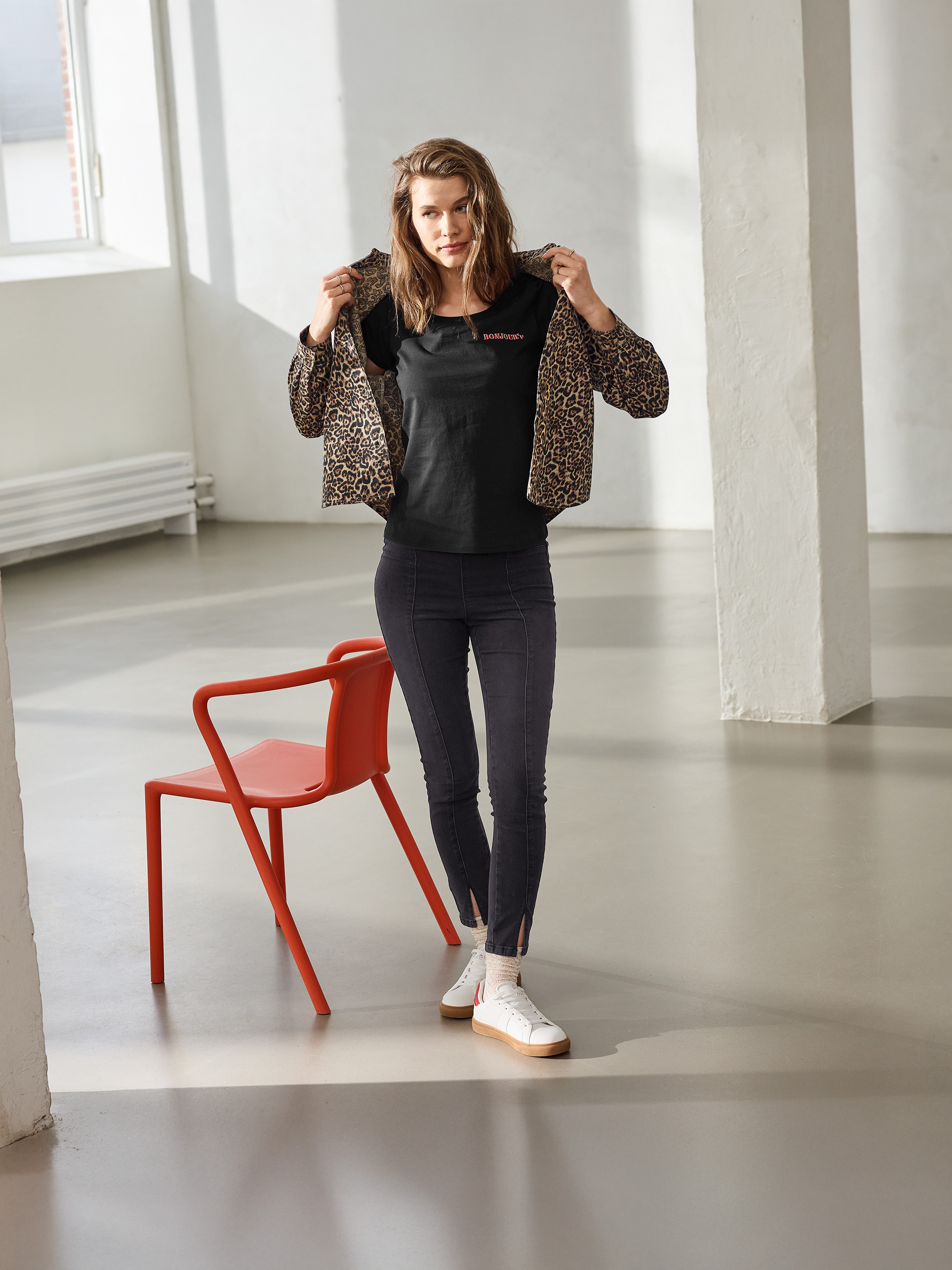 Vrouw in luipaardjasje, zwart 'BONJOUR' T-shirt en zwarte skinny jeans, poserend naast een rode stoel.