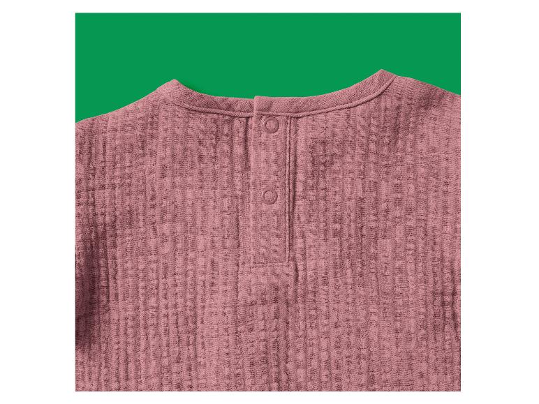 Roze mousseline blouse met knopen in de nek, tegen een groene achtergrond.