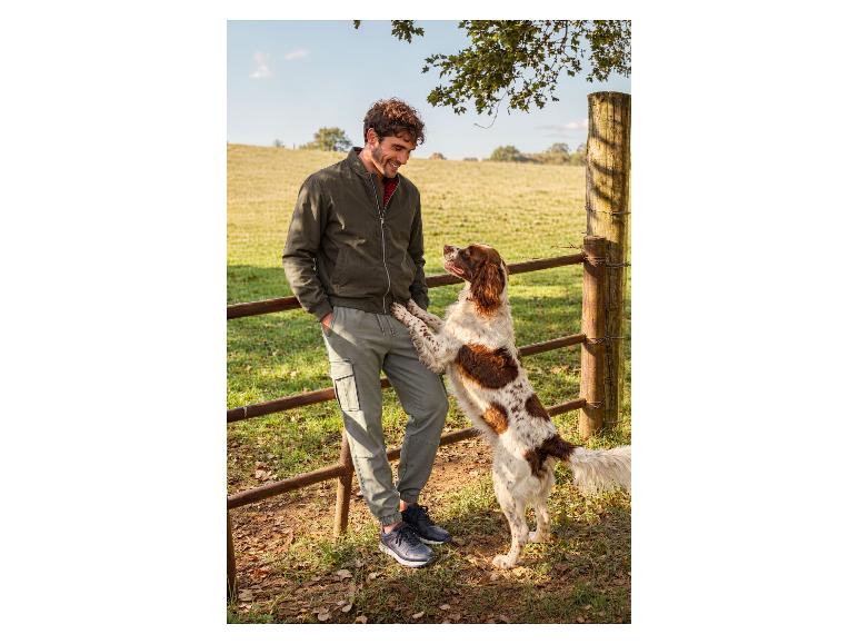 Man in olijfgroene jas en grijze broek met hond op het platteland.
