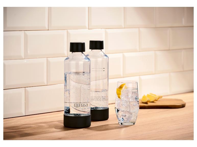 Twee doorzichtige flessen bruiswater met zwarte doppen op een houten aanrecht, naast een glas ijswater met citroen.